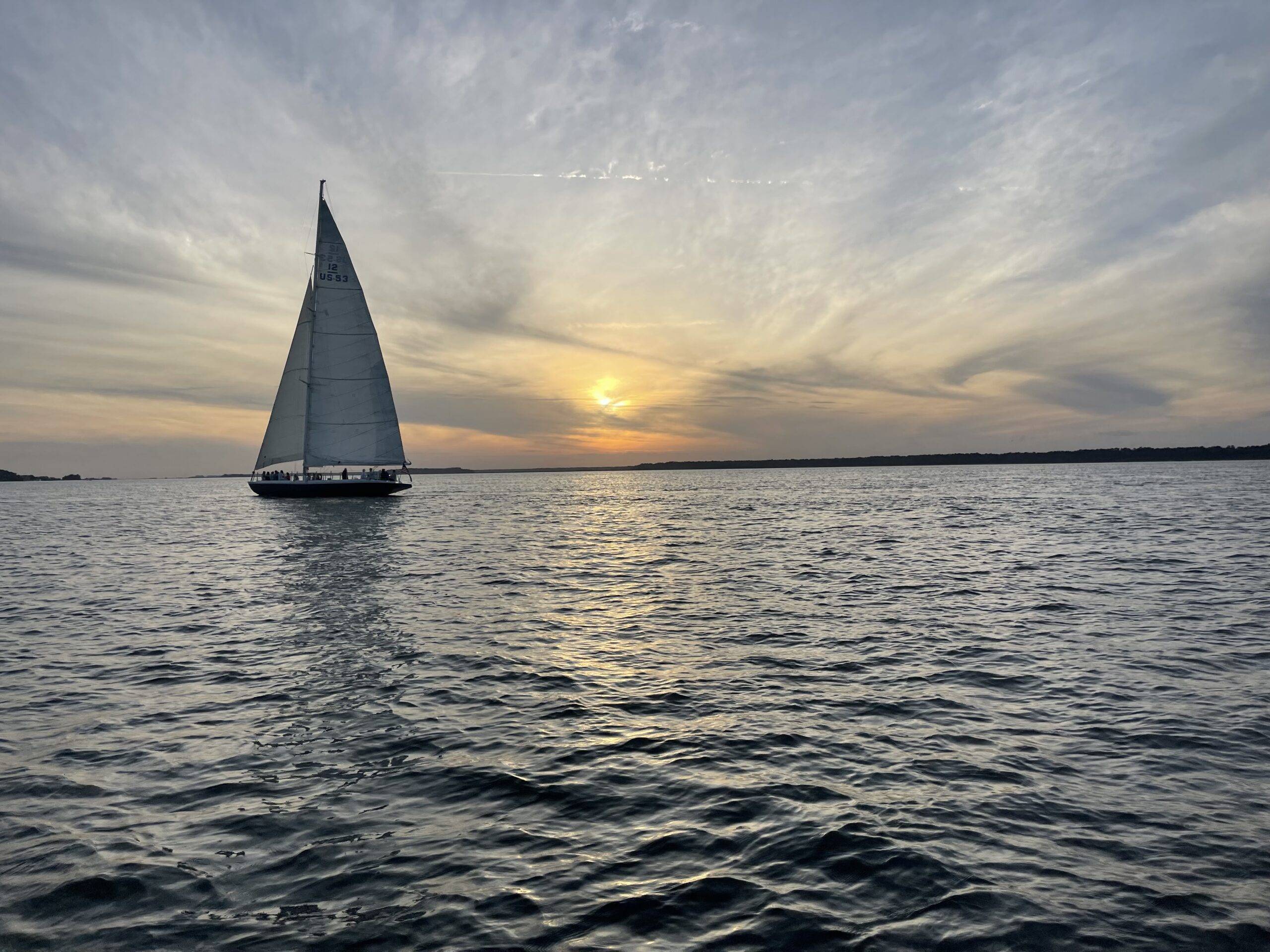 A sailboat glides across the water, illuminated by the warm hues of a sunset in the background.